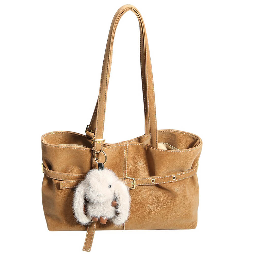 A tan faux-leather tote bag with dual shoulder straps, a front strap detail with gold-tone rivets and buckle, and a white fluffy bunny plush keychain hanging from a ring.